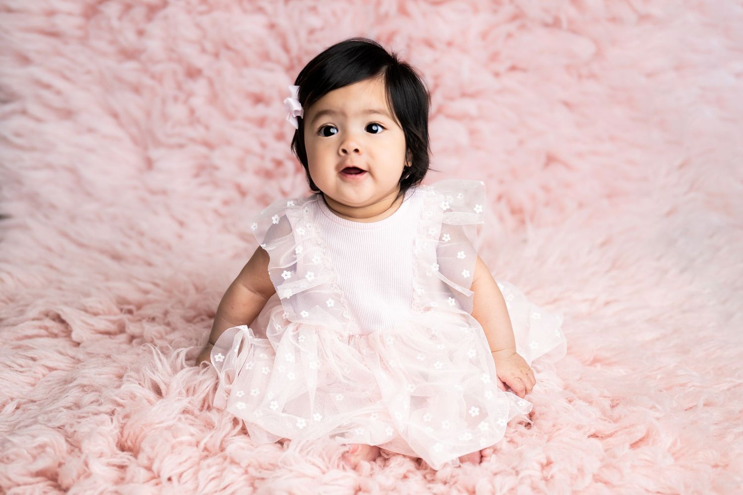 Baby in white lace dress sitting on pink furry blanket during portrait session.