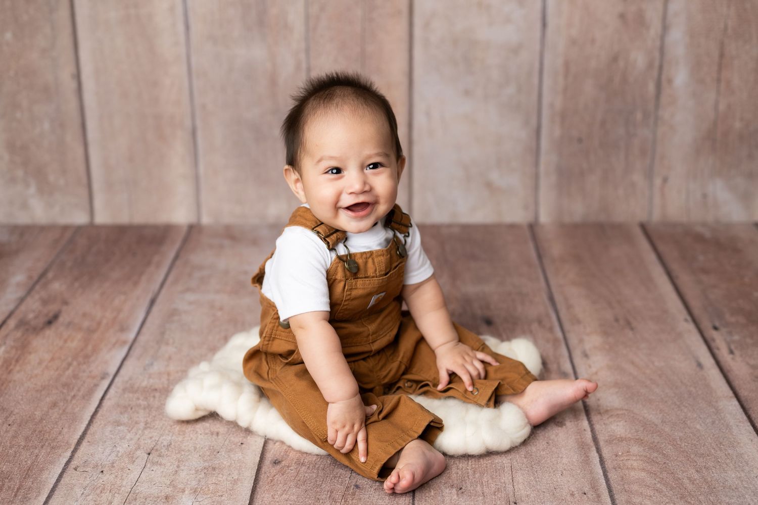 Happy baby wearing brown corduroy overalls sits on white fur blanket against wooden plank backdrop.