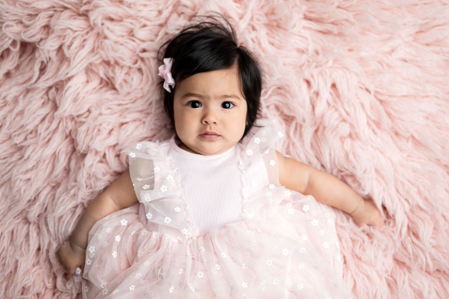 Overhead view of baby wearing white dress lying on pink textured backdrop.