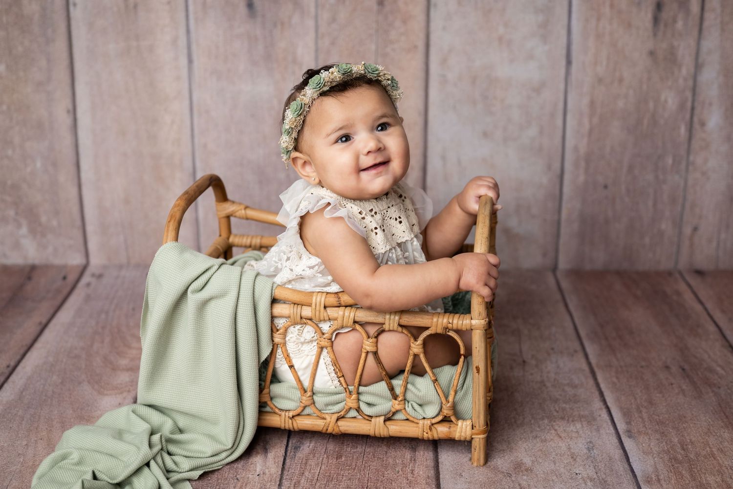 Baby sitting in vintage wicker doll bed wearing white lace dress with mint green blanket against wooden backdrop.