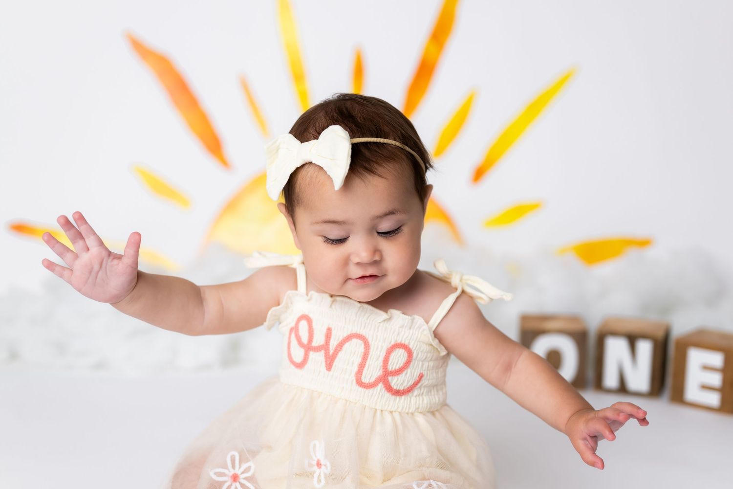 Baby in cream tulle dress celebrating first birthday with hand drawn yellow sunburst backdrop and ONE blocks.