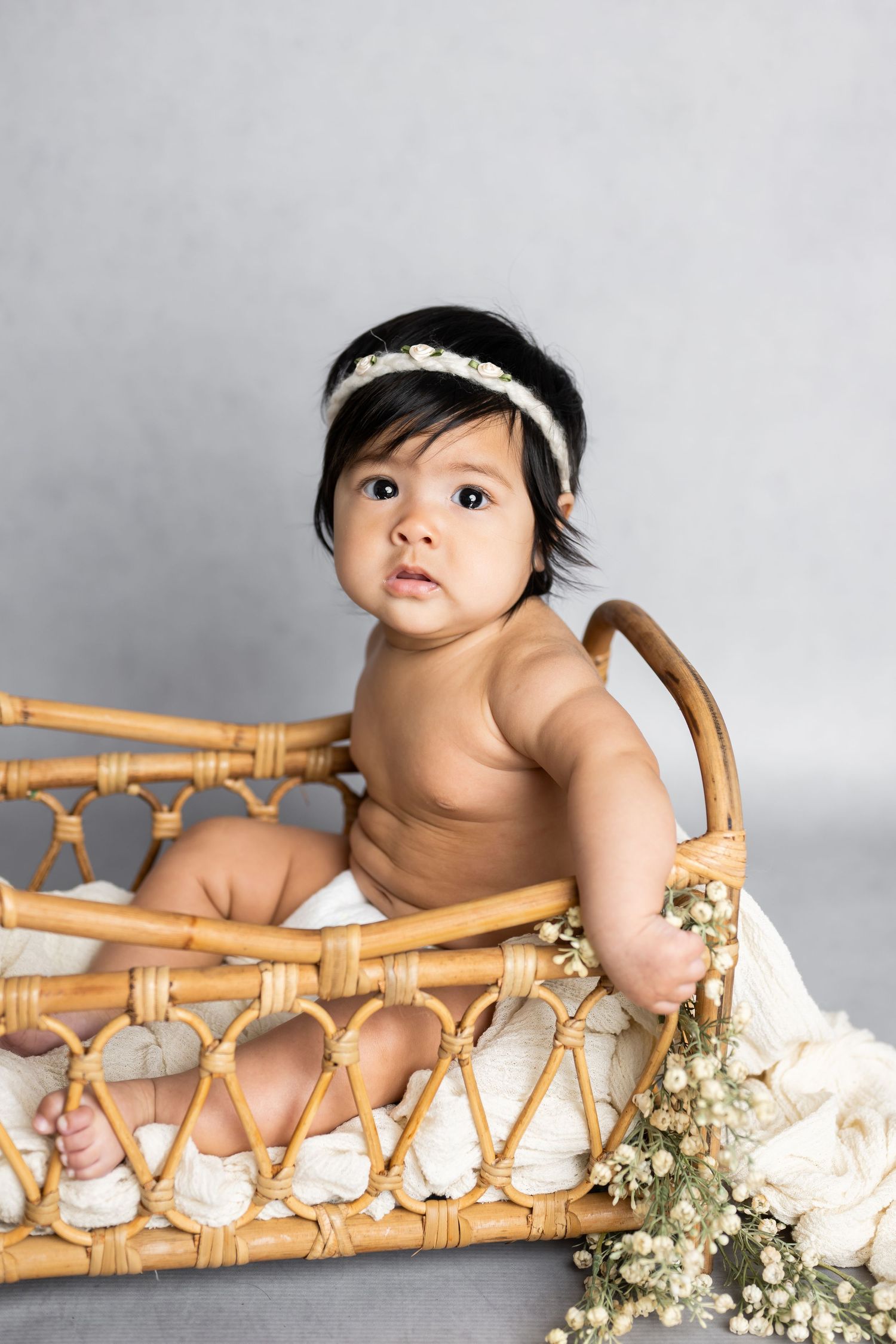 Baby with headband sitting in vintage rattan basket with flowers.
