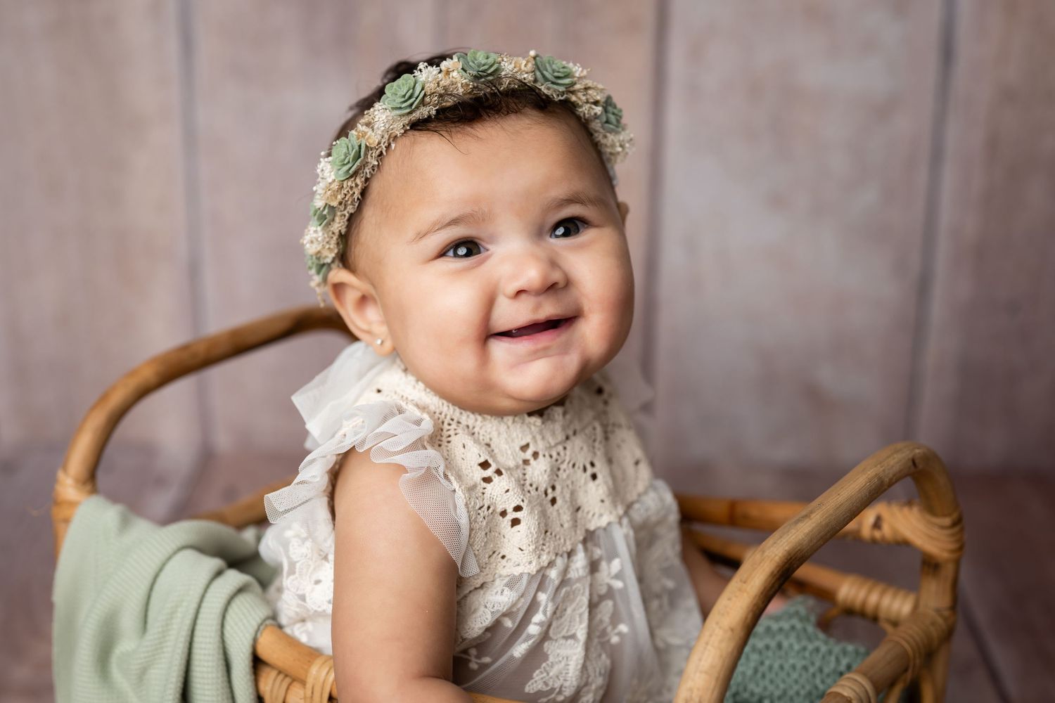Happy baby wearing a white lace dress and floral crown sits in a wooden chair against brown backdrop.