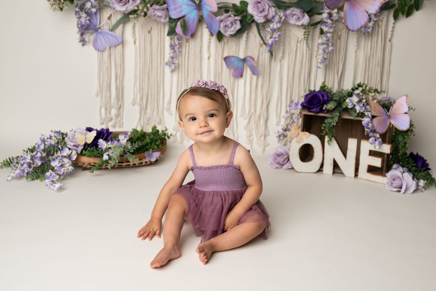 Purple floral themed first birthday setup with macrame backdrop and 'ONE' sign.
