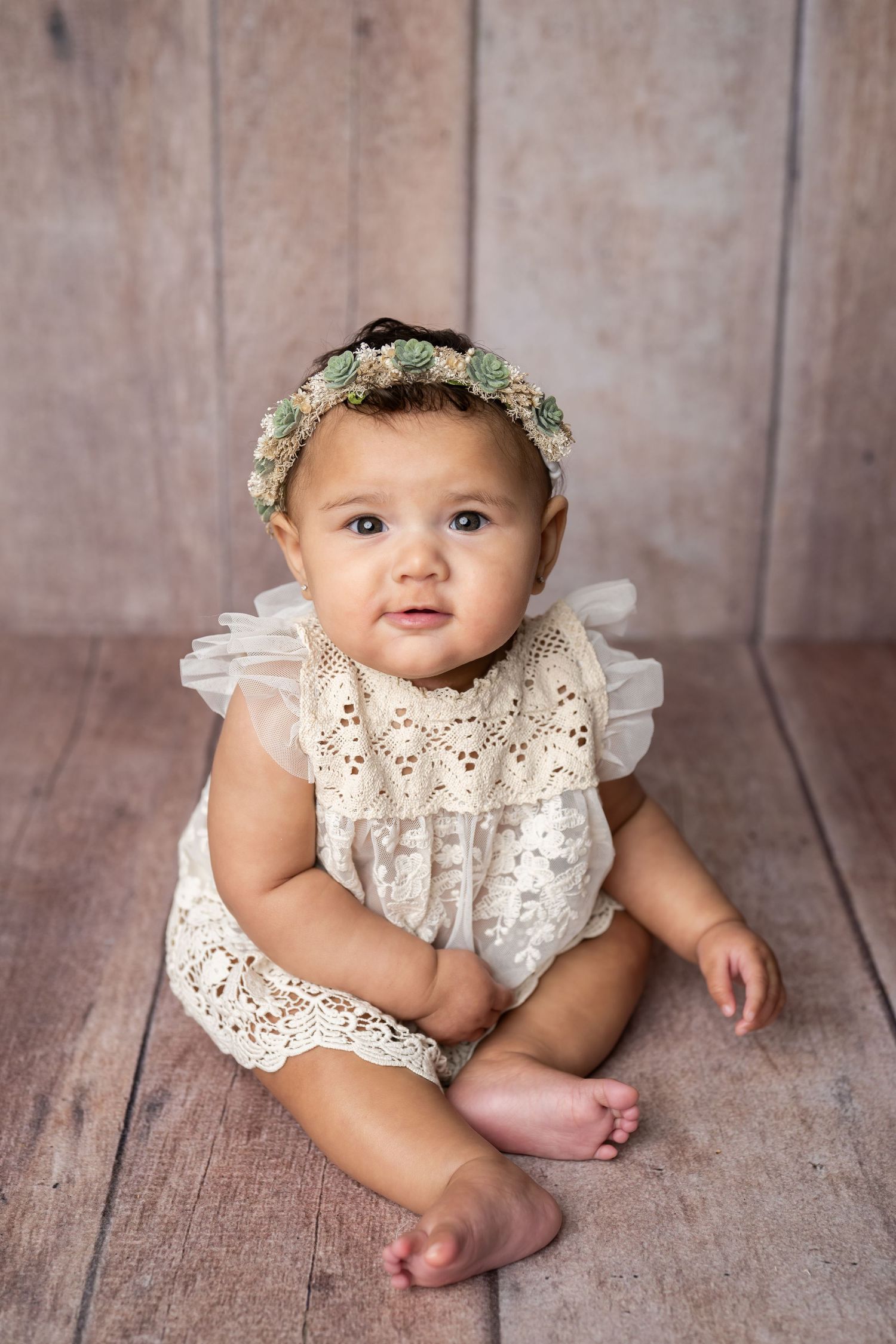 Baby wearing white lace romper and floral crown sits on wooden floor background in vintage-style portrait.