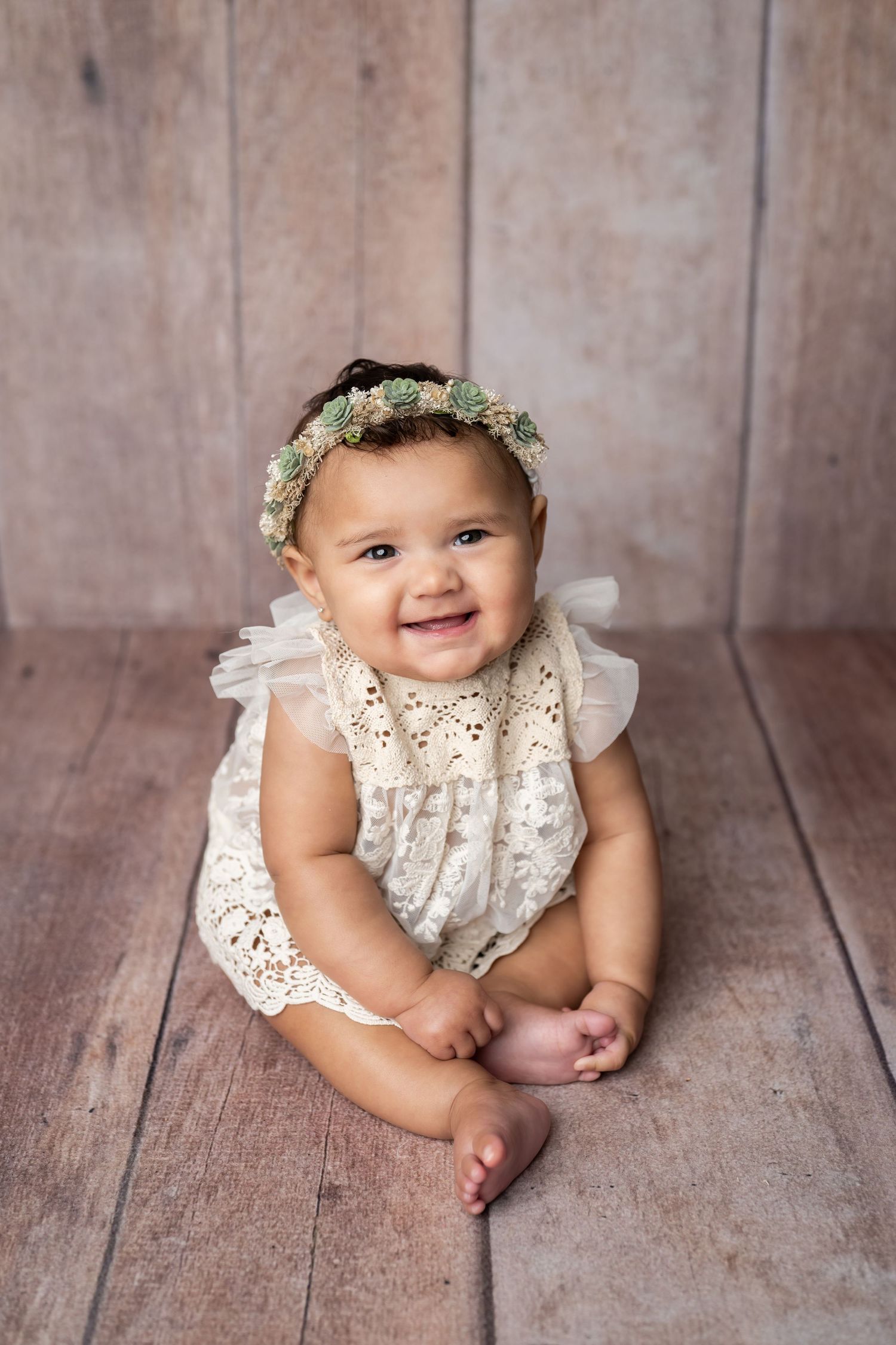 Baby wearing white lace romper and floral crown sits smiling on wooden floor during portrait session.
