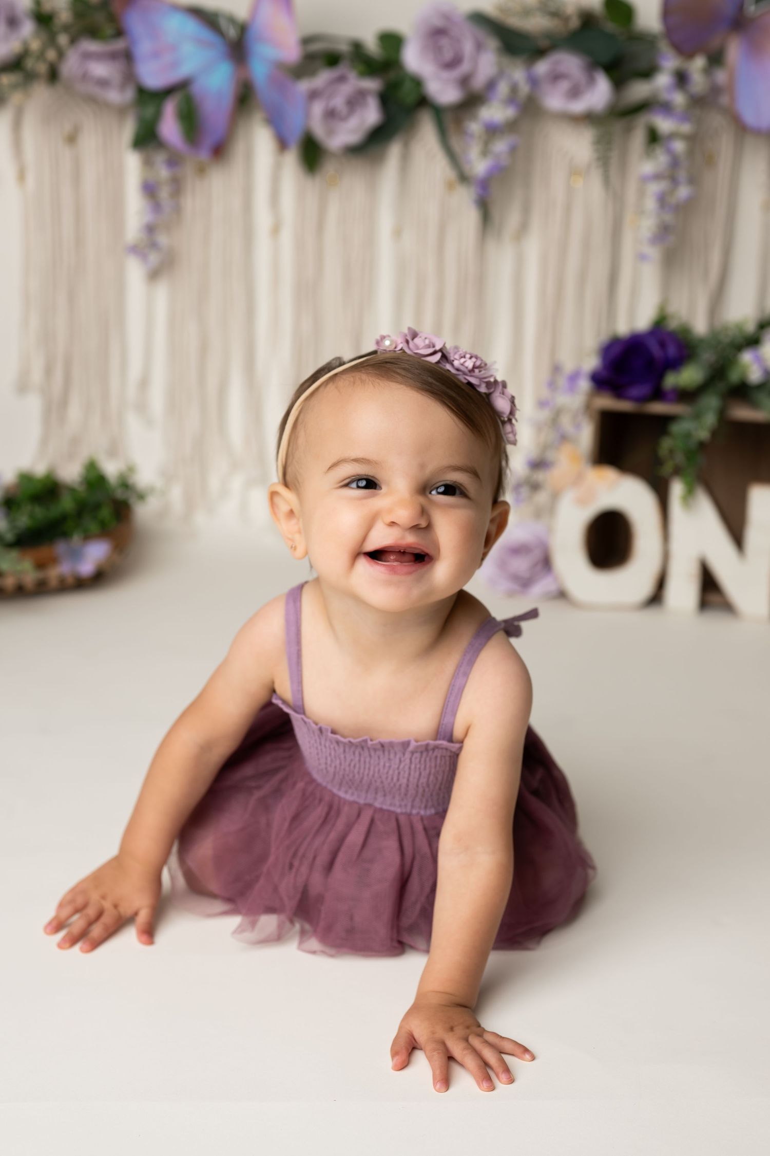 Smiling first birthday portrait with purple dress and floral backdrop decorations.