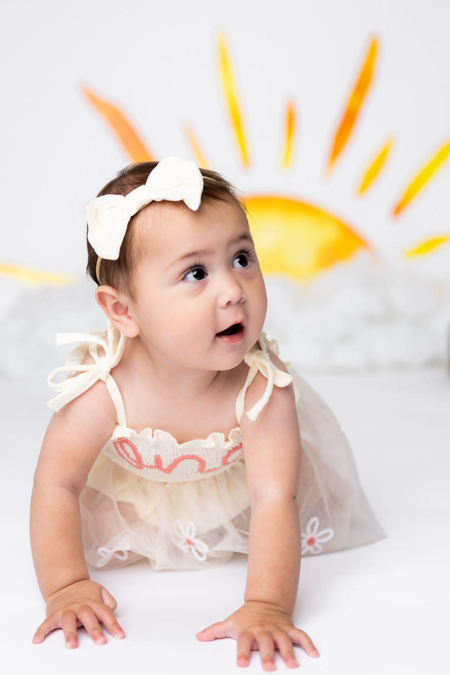 Baby in white dress and bow crawls on white floor with painted yellow sun backdrop.