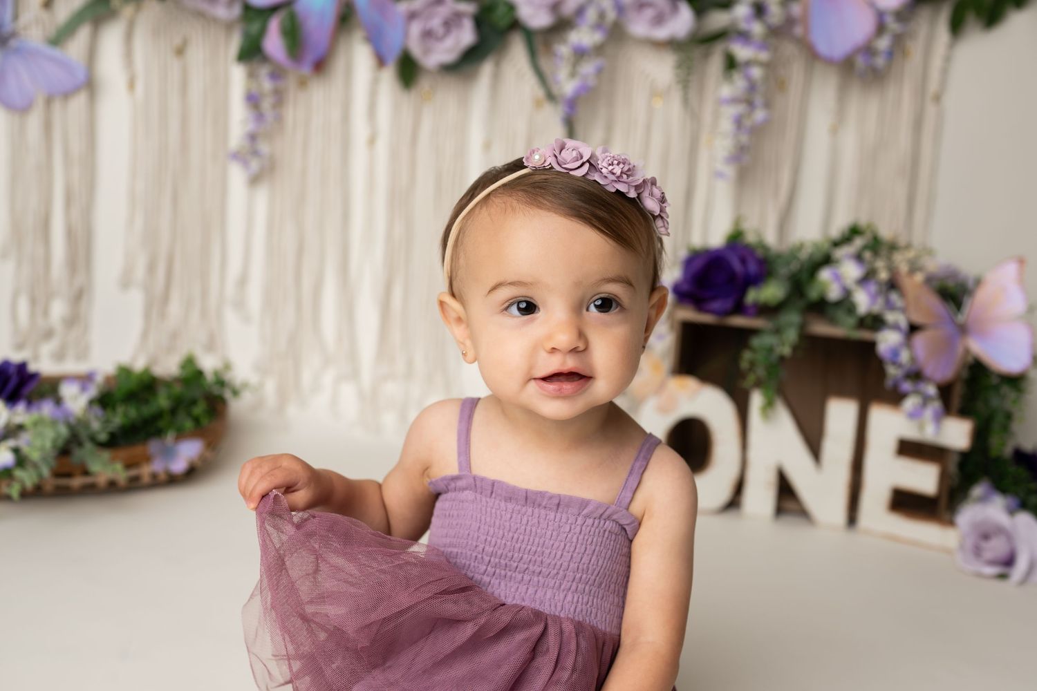 A first birthday photo featuring purple decor, butterflies, and 'ONE' text against a cream backdrop.