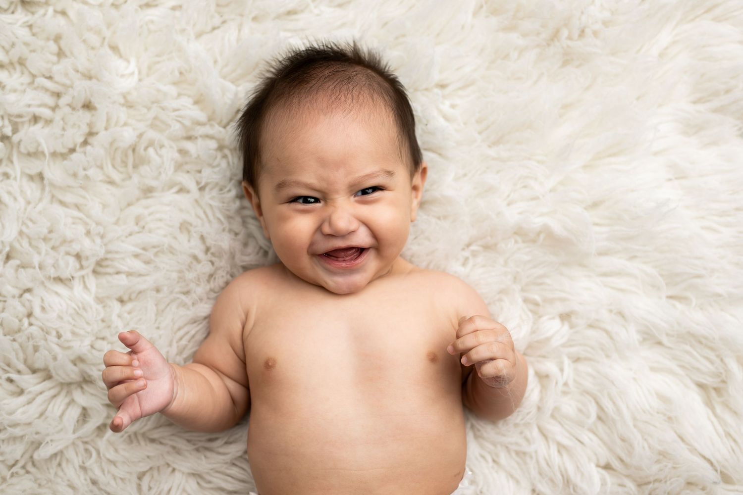 Happy baby lying on soft white fur blanket with a big gummy smile and bare chest.