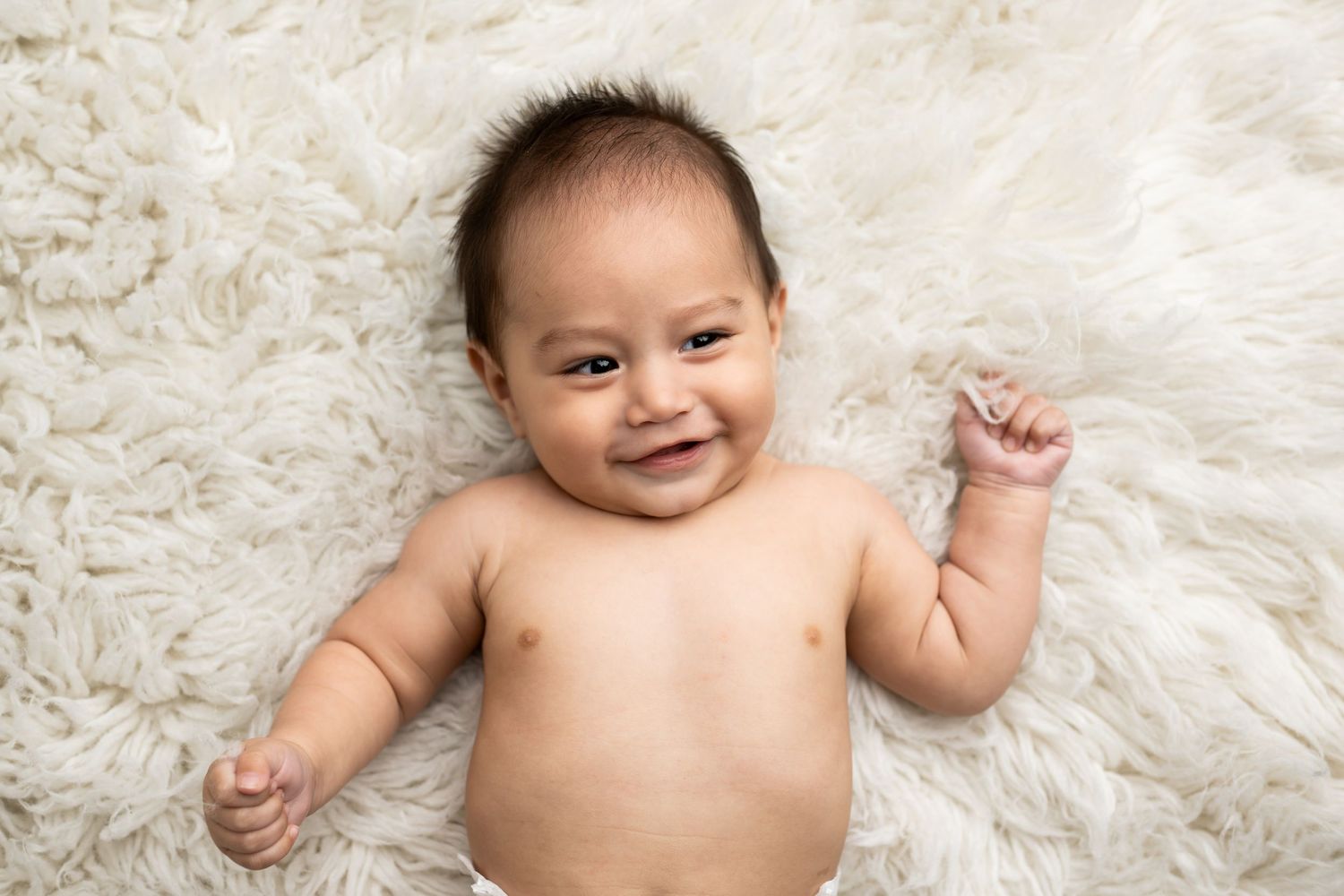 Smiling baby lying on soft white fur blanket with raised arms.