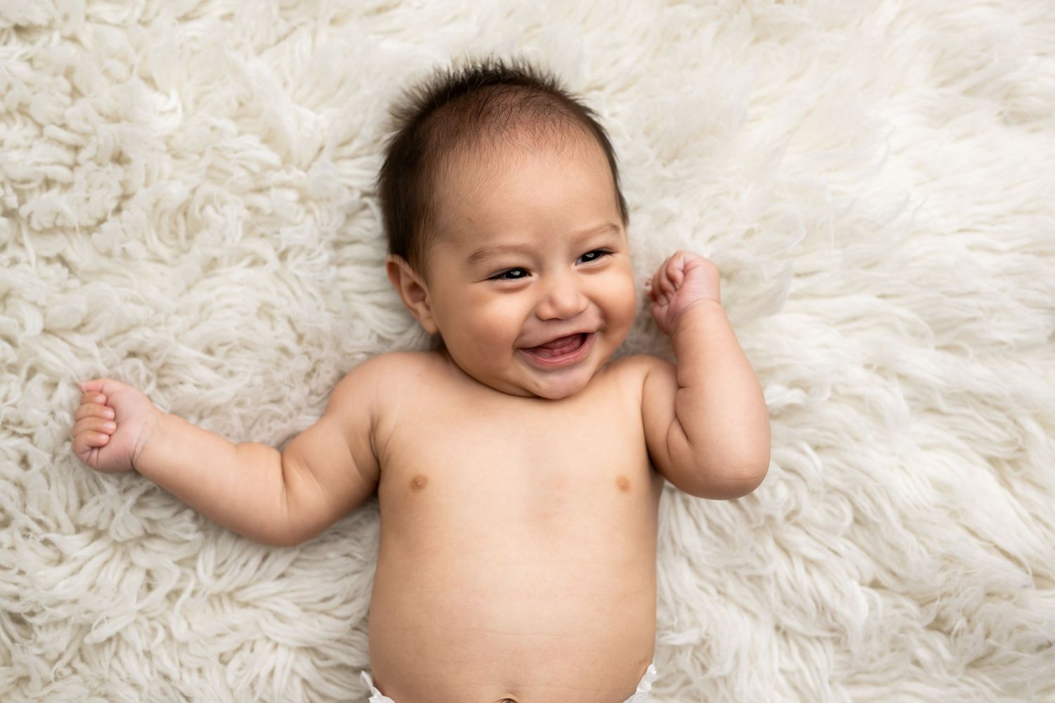 Happy bare-chested baby lying on plush white fur blanket smiling joyfully with arms outstretched.