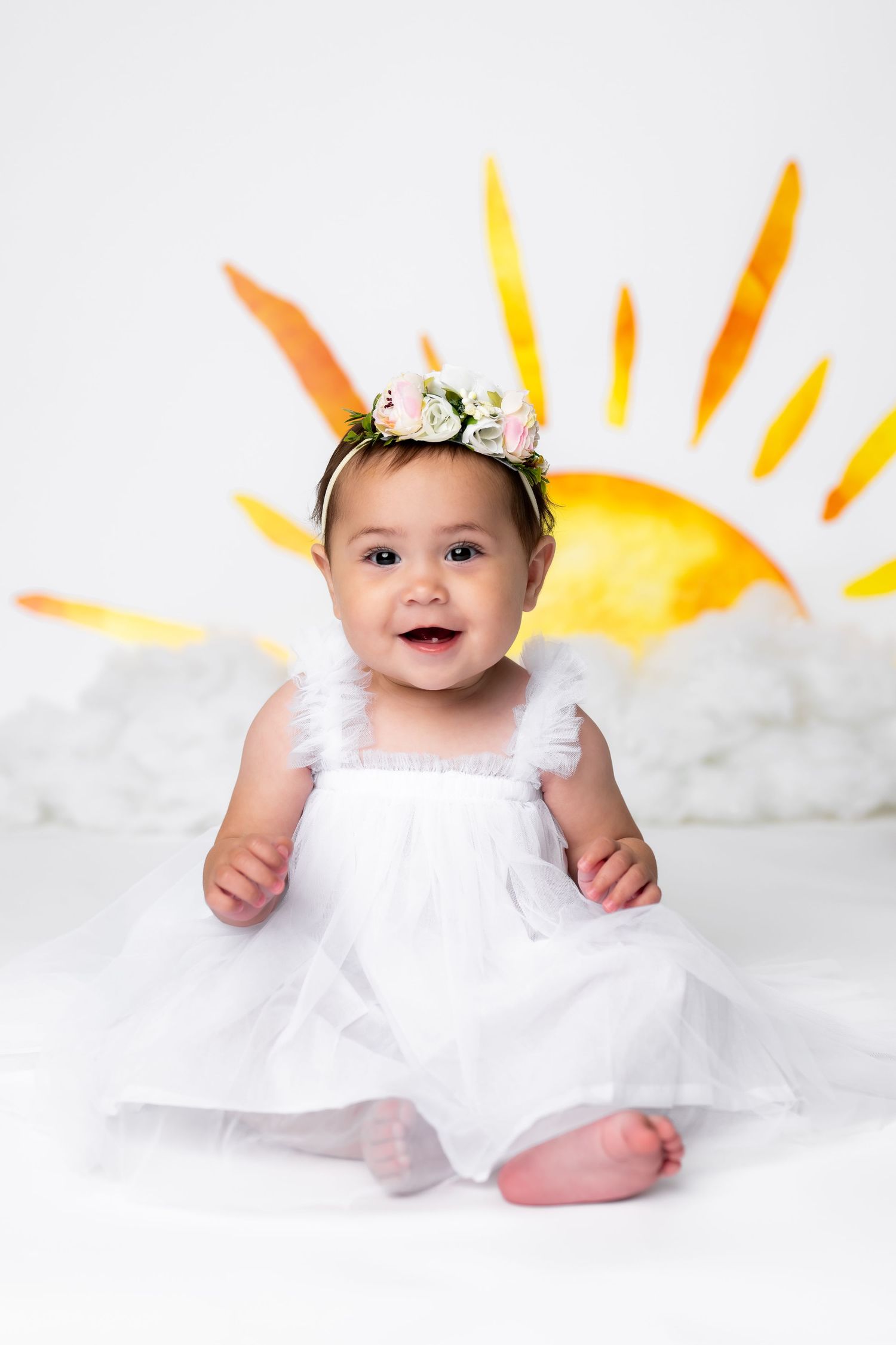 A small baby wearing a white dress and floral headband sits in front of a painted sunshine backdrop.