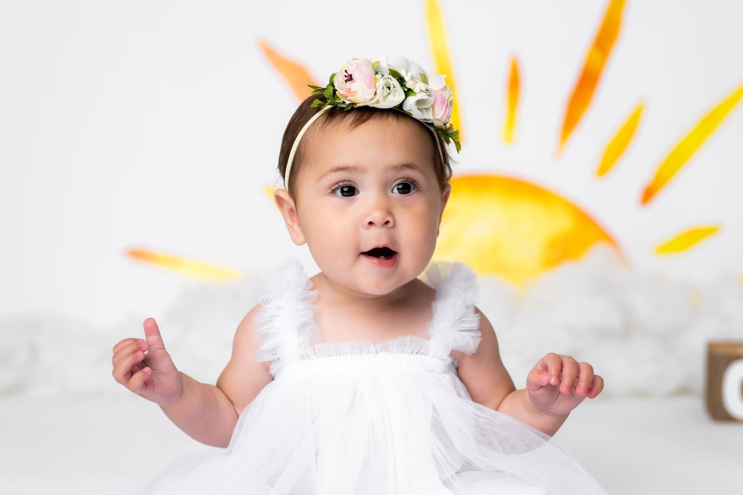 Baby in white dress wearing floral crown poses against painted sun backdrop for whimsical photo.