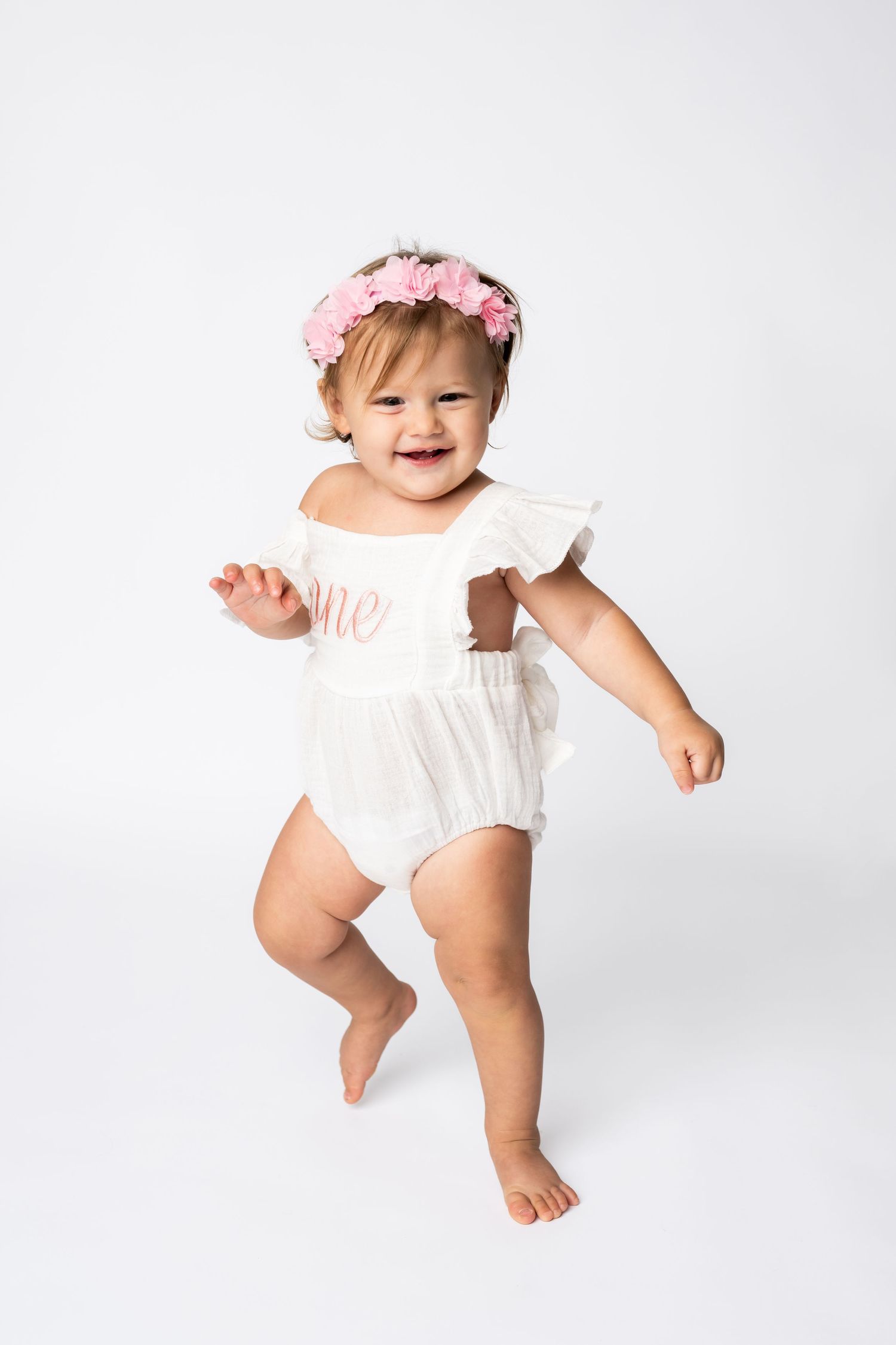 A joyful portrait in white romper with pink floral crown against white background.