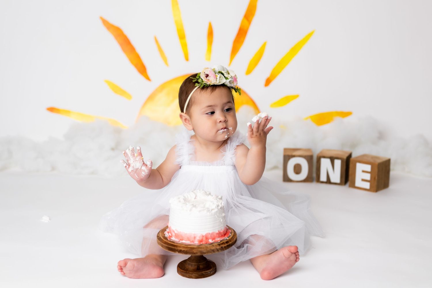 Baby in white dress sits near wooden blocks and cake with hand-drawn yellow sunshine backdrop during first birthday photoshoot.