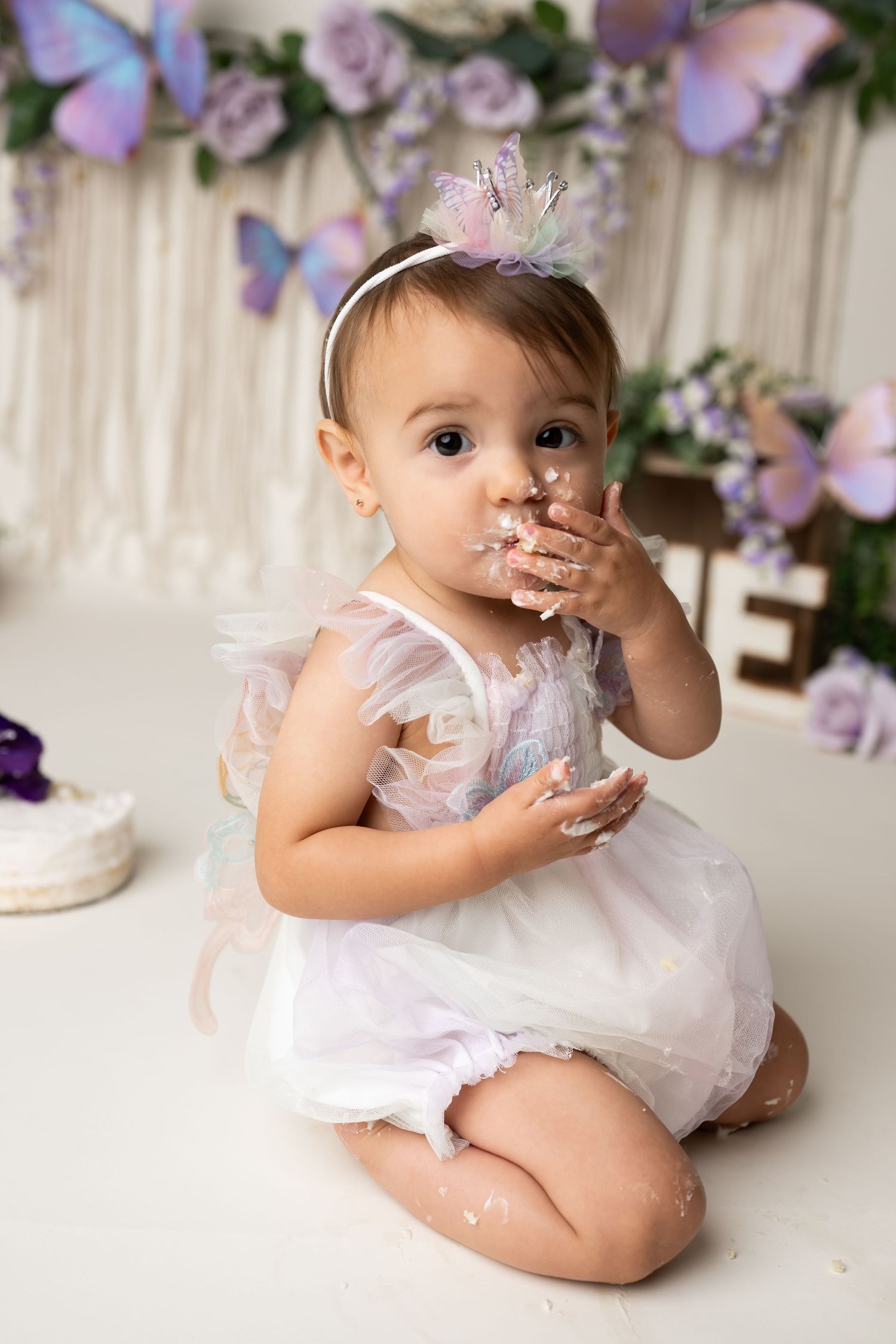 A messy cake smash photo with white dress and purple butterfly backdrop.