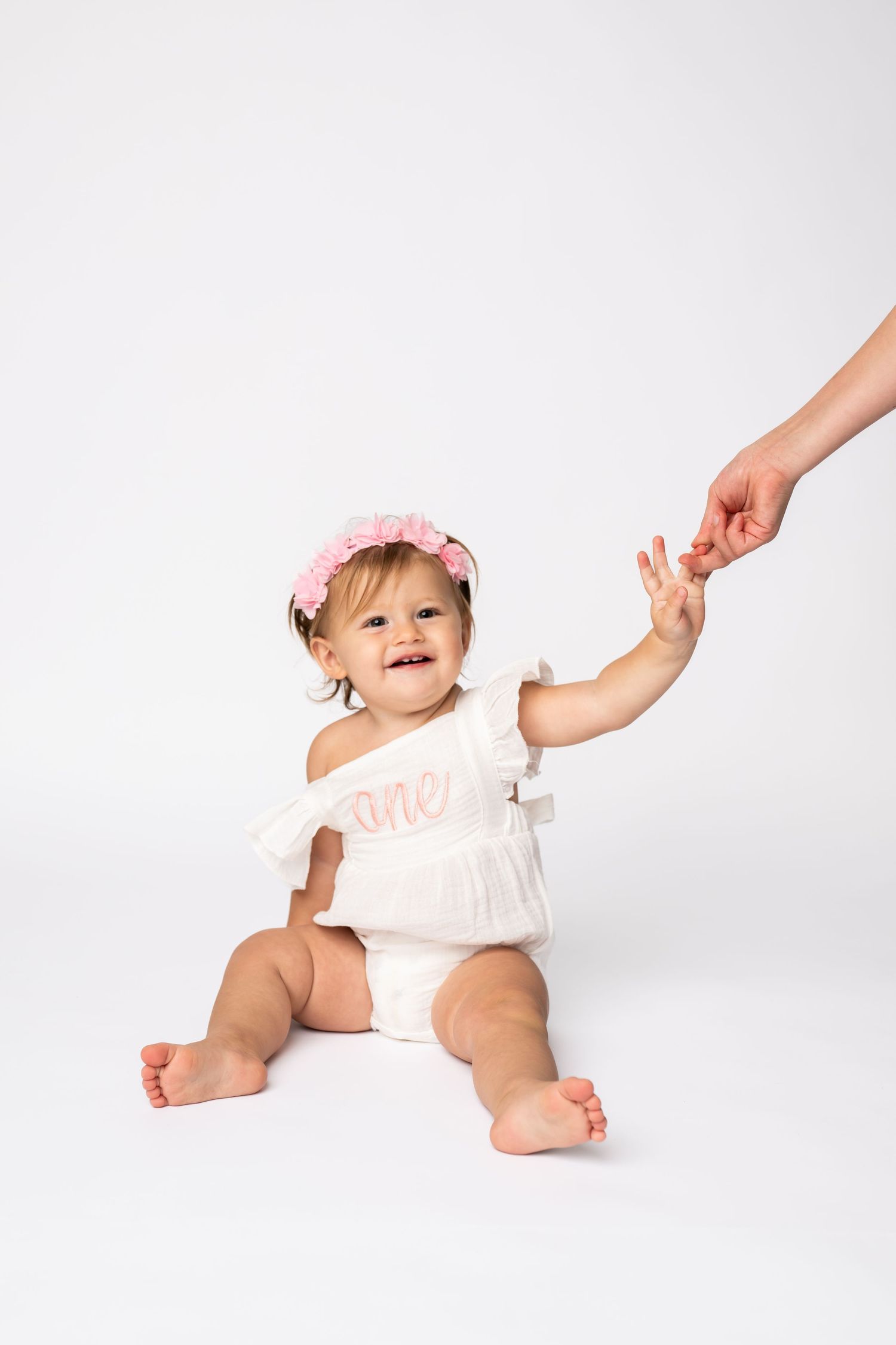 A celebratory pose in white outfit with pink floral crown against studio backdrop.