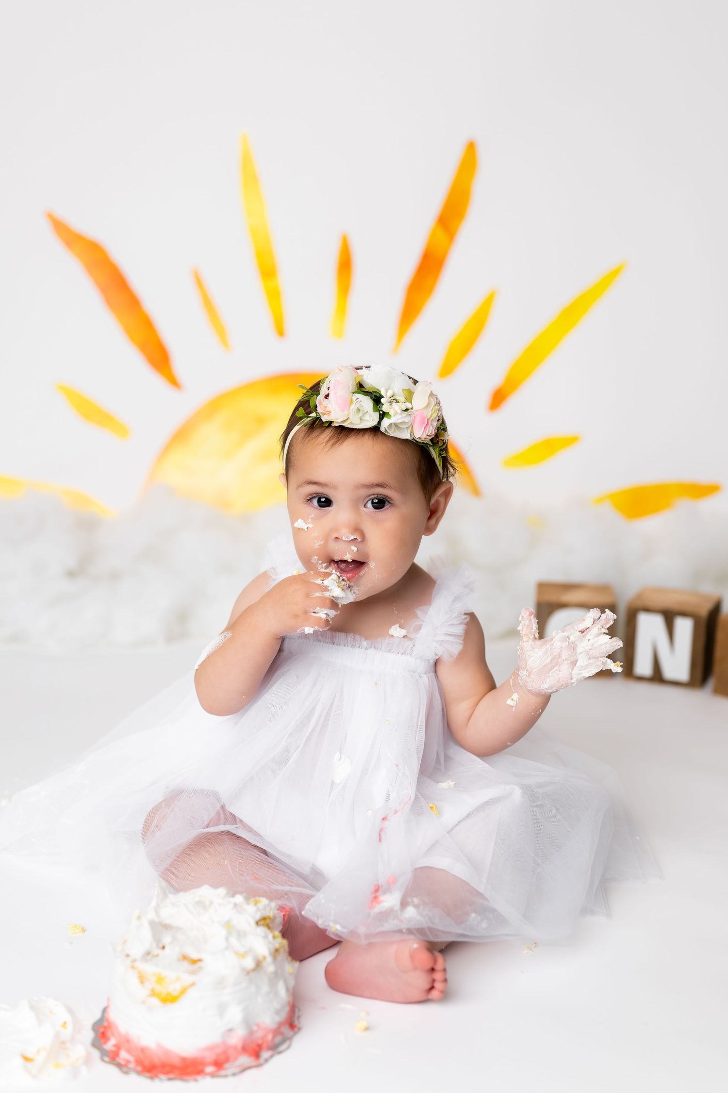 A baby in white enjoying their first birthday cake smash photo session with a yellow sun backdrop.