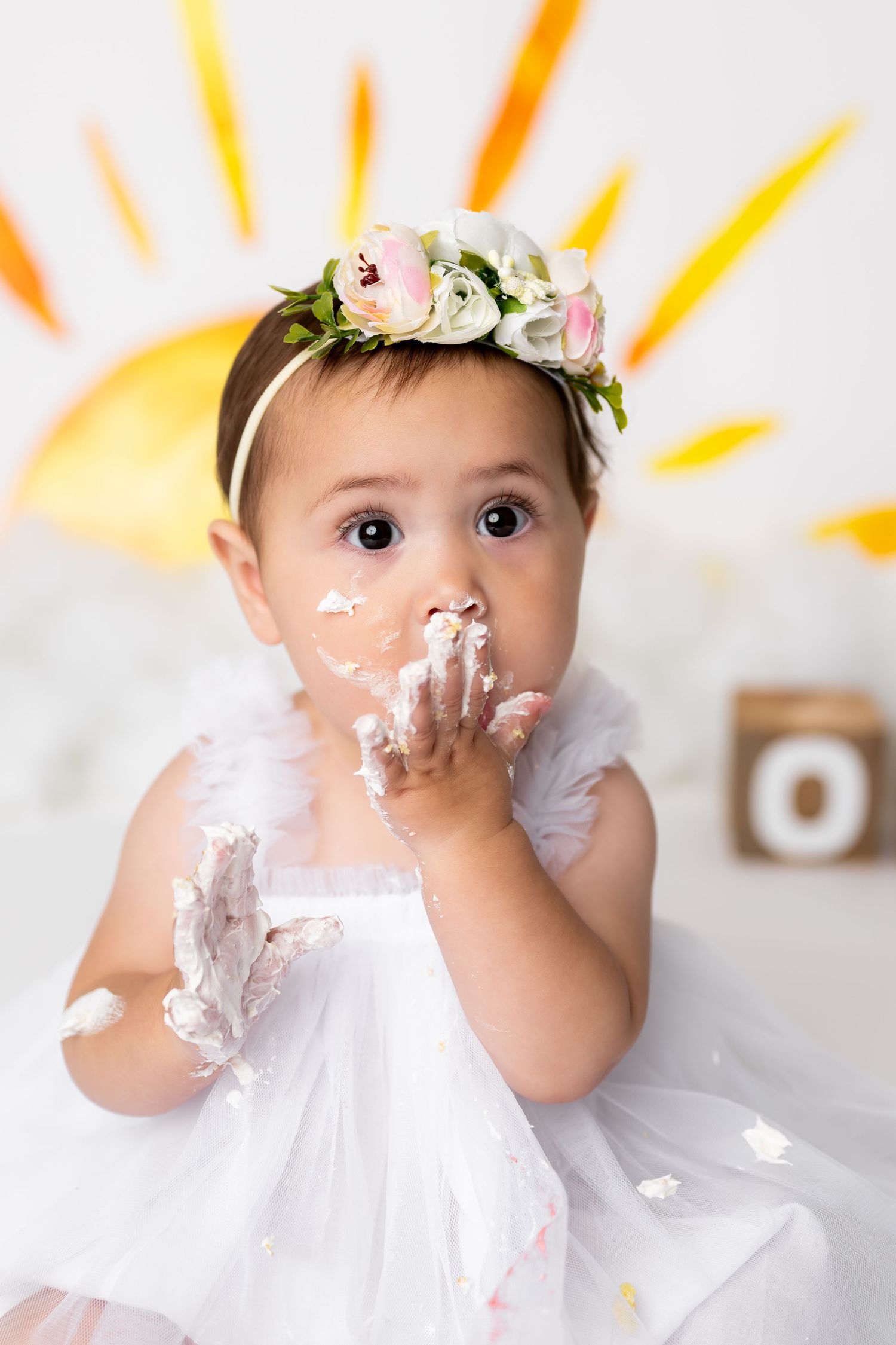 Baby wearing white dress and floral crown gets messy with cake during first birthday photoshoot against yellow sunburst backdrop.