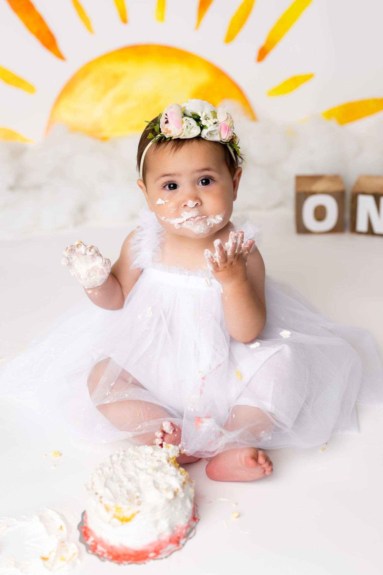 Baby in white tutu dress and floral crown enjoys first birthday cake smash photoshoot with painted sunrise backdrop.