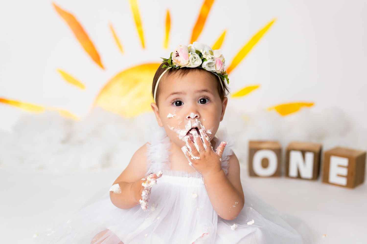 A toddler in a white tulle dress and crown sits with wooden letter blocks spelling ONE behind them in a birthday photoshoot.