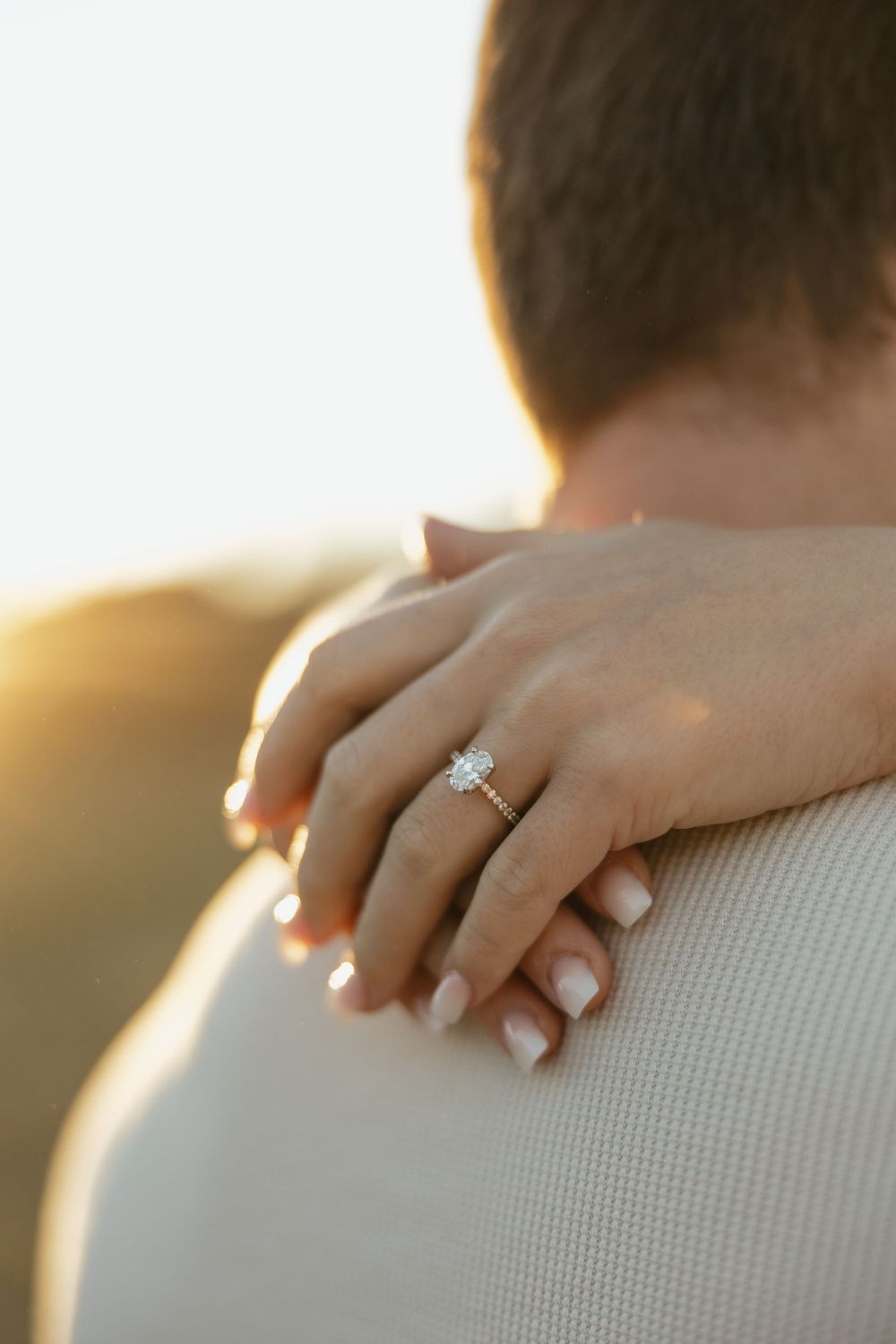 Close-up of an engagement ring and manicured hand against a shoulder during golden hour.