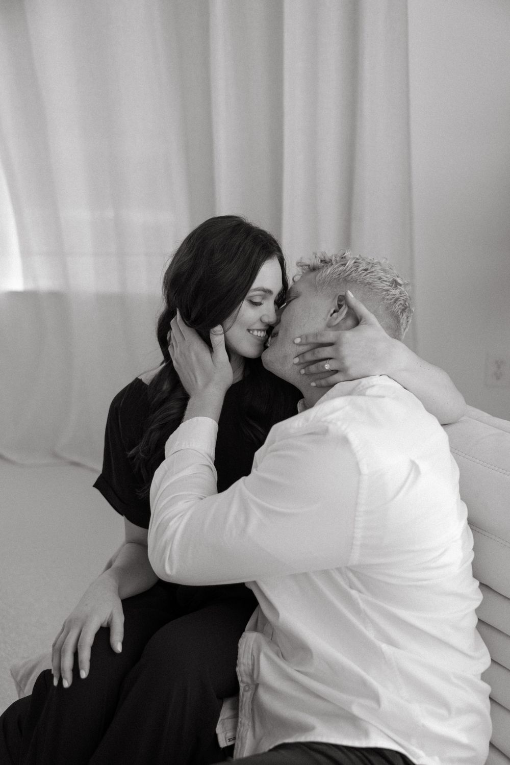 An intimate black and white portrait of a couple sharing a tender moment against white curtains.