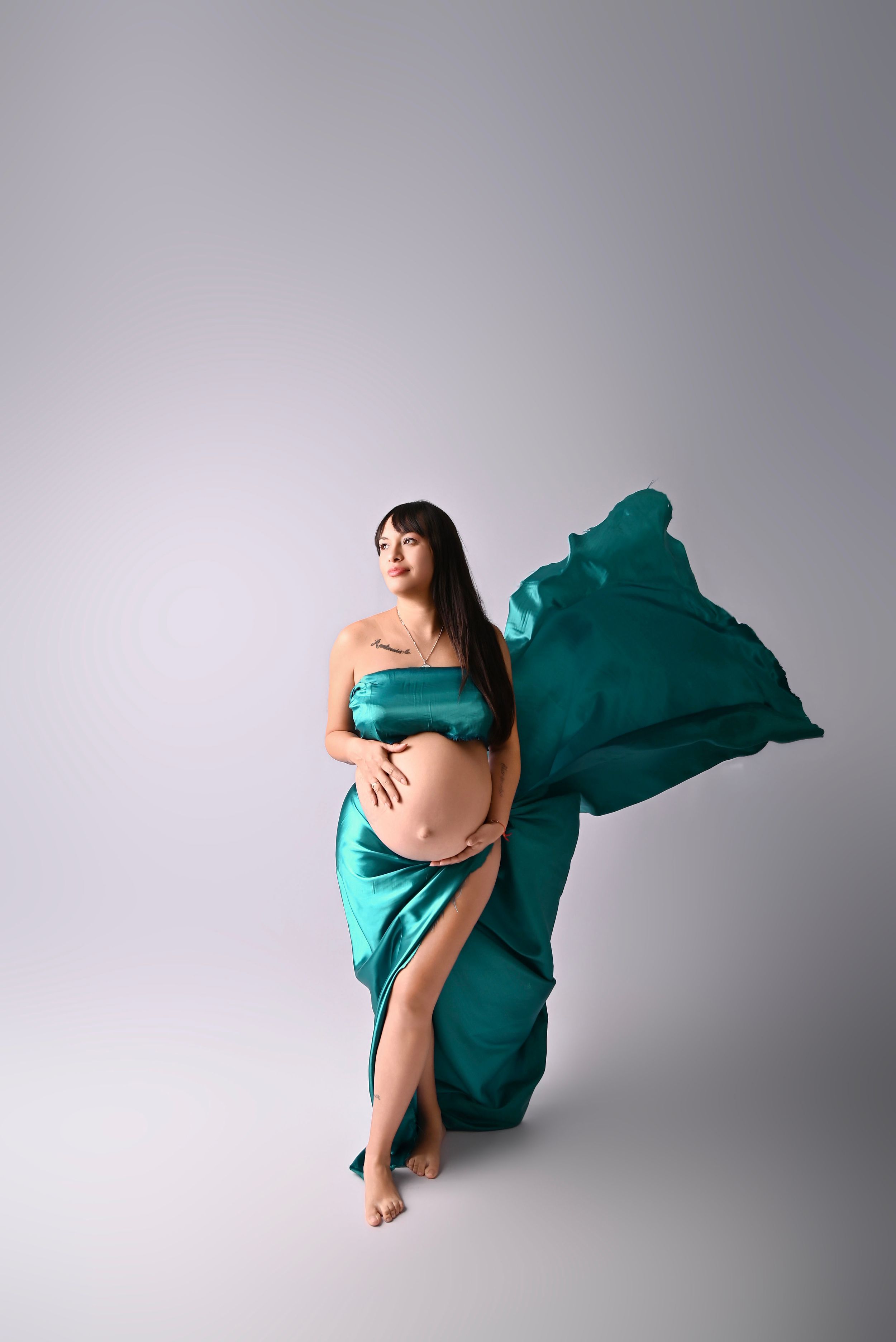 Pregnant woman draped in flowing teal silk fabric against gray backdrop, cradling baby bump in elegant maternity portrait.