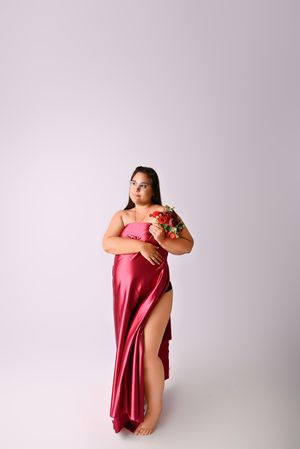 Woman in red satin slip dress holding floral bouquet, posing barefoot against white background.
