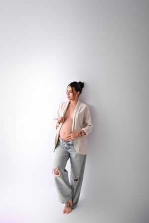 Pregnant woman in open cream blazer and ripped jeans leaning against white wall, showcasing baby bump.