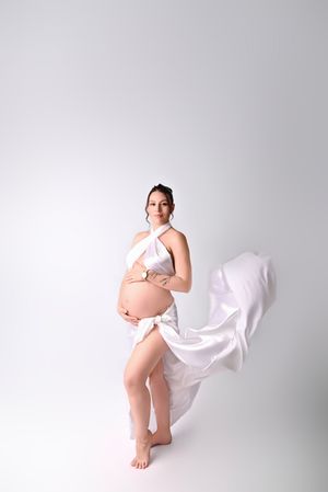 Pregnant woman posing in flowing white fabric wrap dress, showcasing baby bump in a bright studio setting.