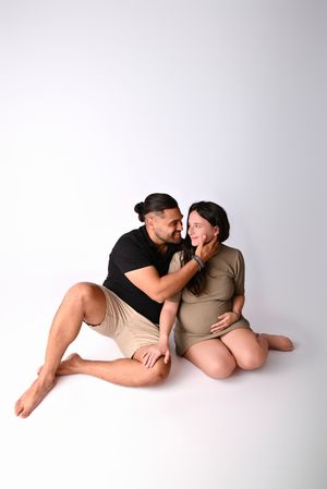 A couple shares a tender moment, with a man gently touching a pregnant woman's face as they sit together on a white background.