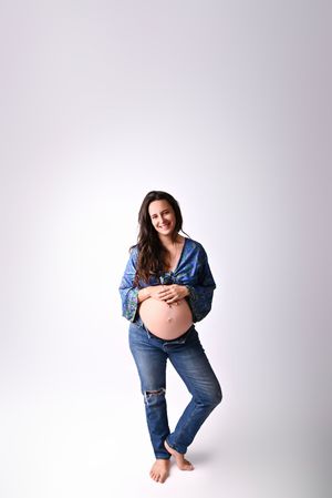 Smiling pregnant woman in blue floral top and ripped jeans cradling her bare baby bump in a studio.