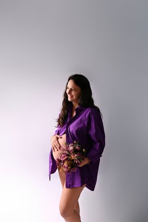 Pregnant woman in purple shirt holding pink flowers, smiling sideways against white background.