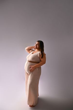 Pregnant woman in a beige maxi dress posing gracefully in a studio, cradling her baby bump with one hand.