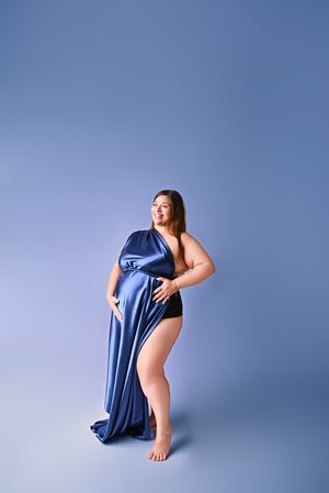 Pregnant woman draped in blue satin fabric posing gracefully against a matching blue studio background.