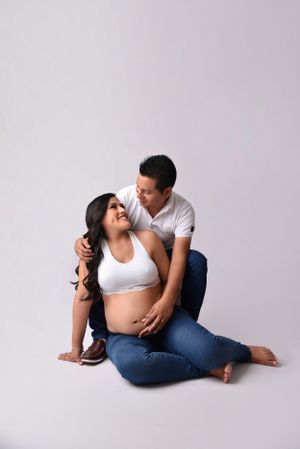 Intimate maternity photoshoot featuring couple sitting together against neutral background.