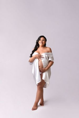 Maternity portrait wearing white off-shoulder dress against light grey background.