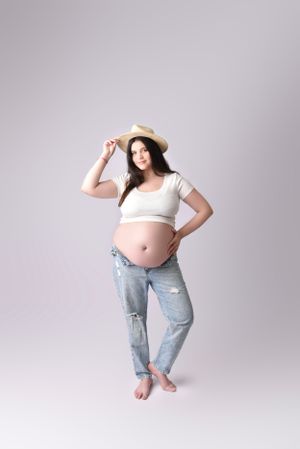 Casual maternity portrait in denim and white crop top with straw hat.
