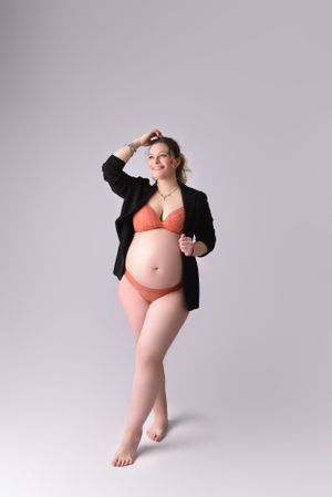 Maternity portrait of woman in black blazer and coral lingerie posing against grey background.