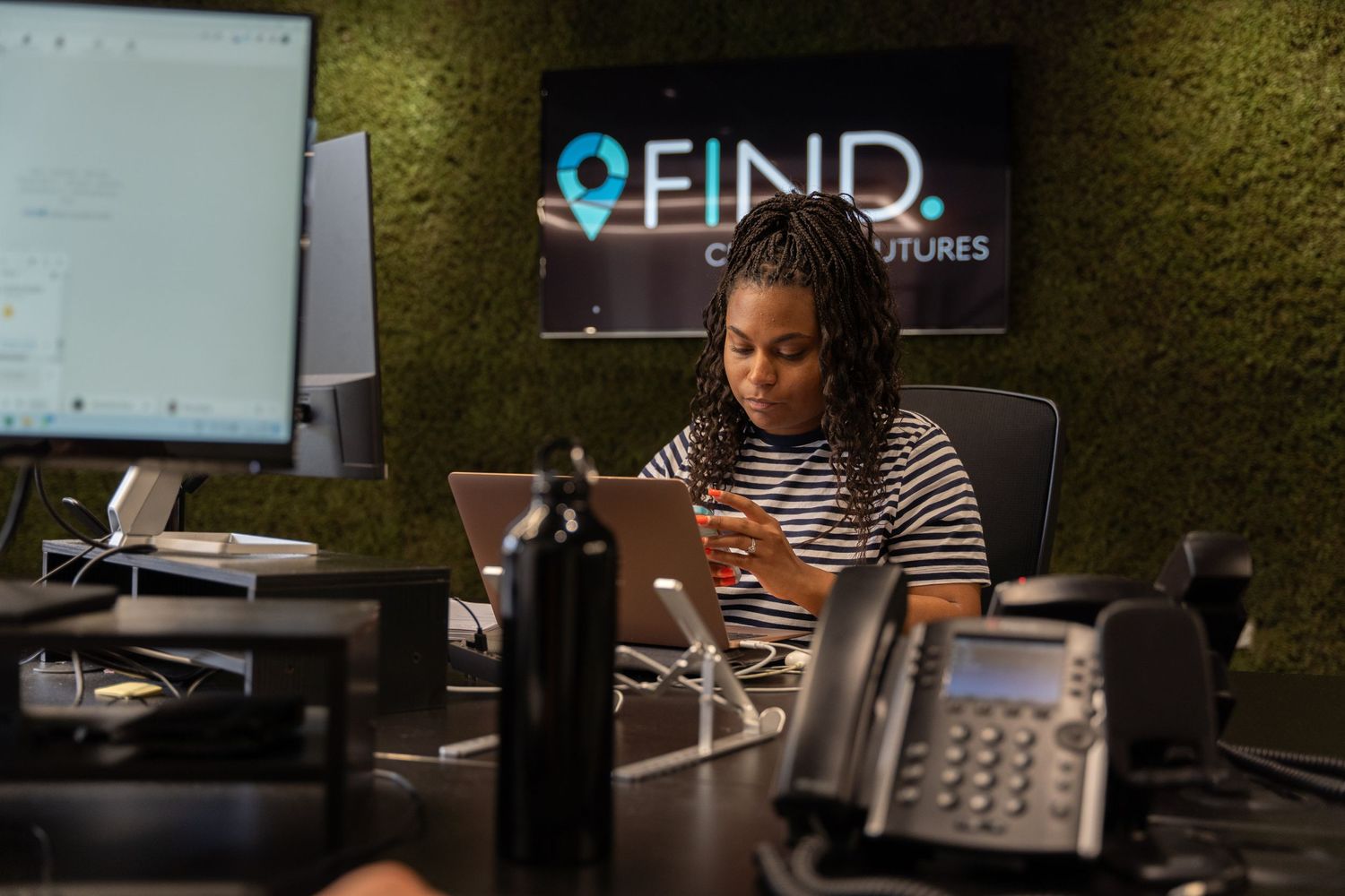 Professional working on laptop at modern office desk with FIND logo visible on wall.