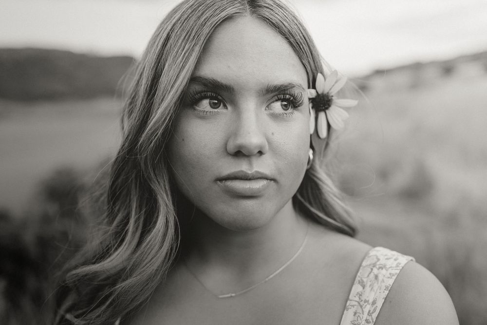 Black and white portrait of a young woman with a daisy in her hair, gazing thoughtfully in a natural outdoor setting.