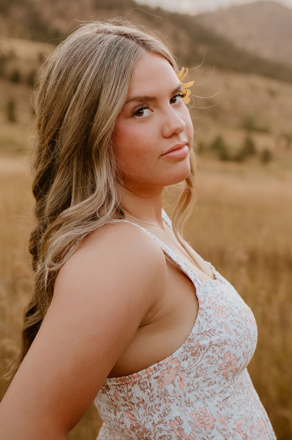 Young woman with blonde hair in a floral dress poses in a golden field with mountains in the background.