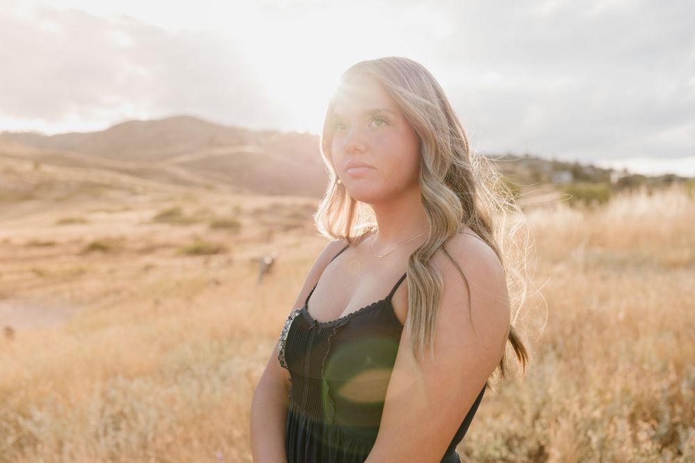 Young woman with blonde hair posing in golden field with sunlit hills in background.