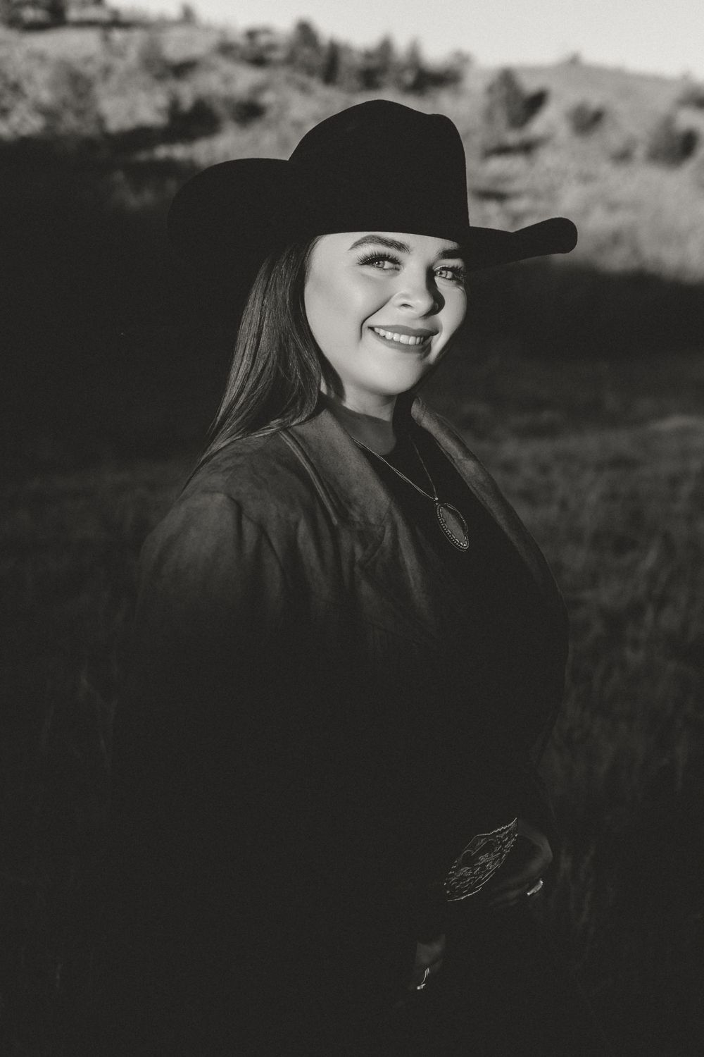 Black and white portrait of a smiling young woman wearing a cowboy hat in a rural outdoor setting.