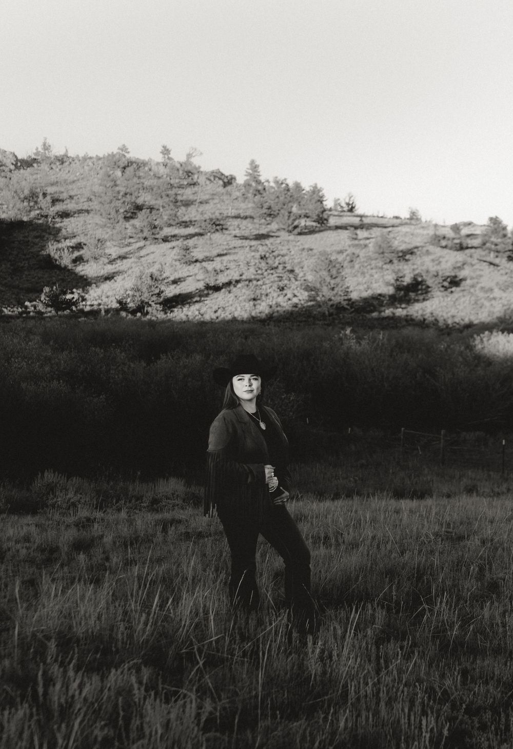 Black and white photo of a person standing in a grassy field with a rocky tree-covered hillside in the background.