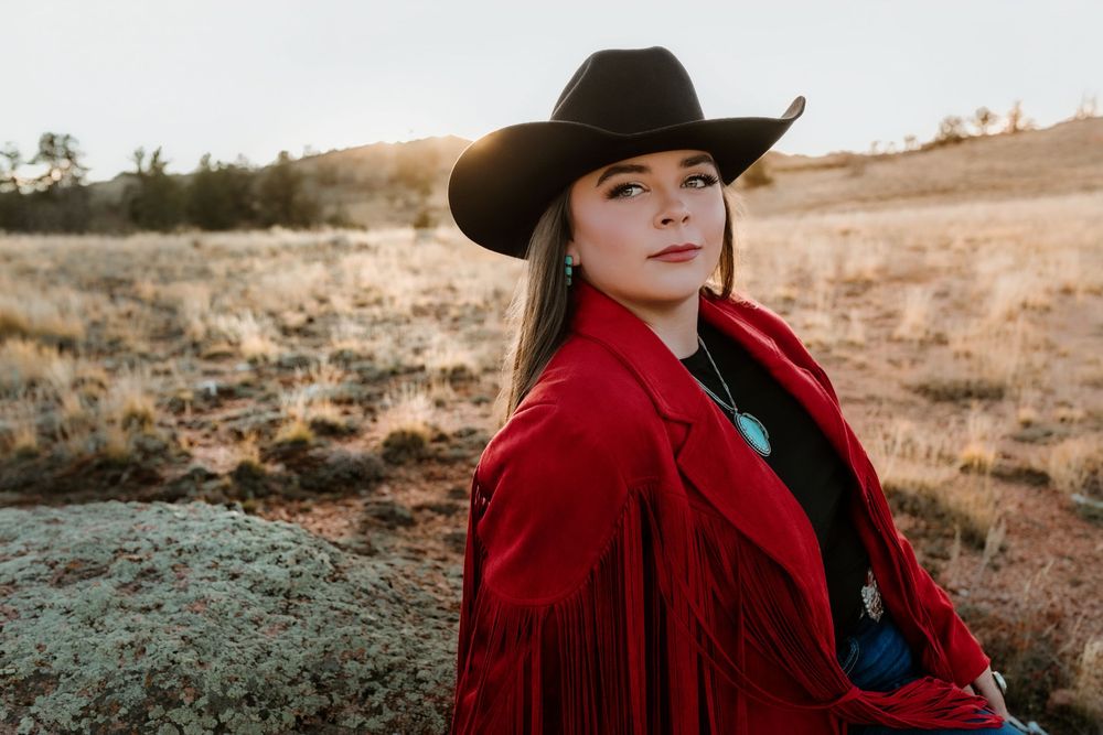Woman in red fringe jacket and black cowboy hat poses in golden field at sunset.