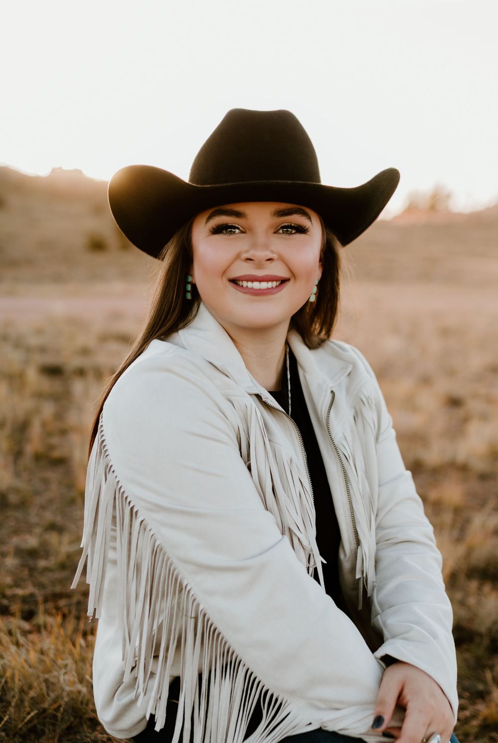 Smiling young woman wearing black cowboy hat and white fringe jacket outdoors at sunset.