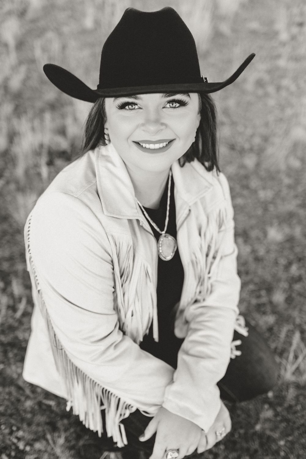 Smiling woman in black cowboy hat and fringed jacket posing outdoors in black and white photo.